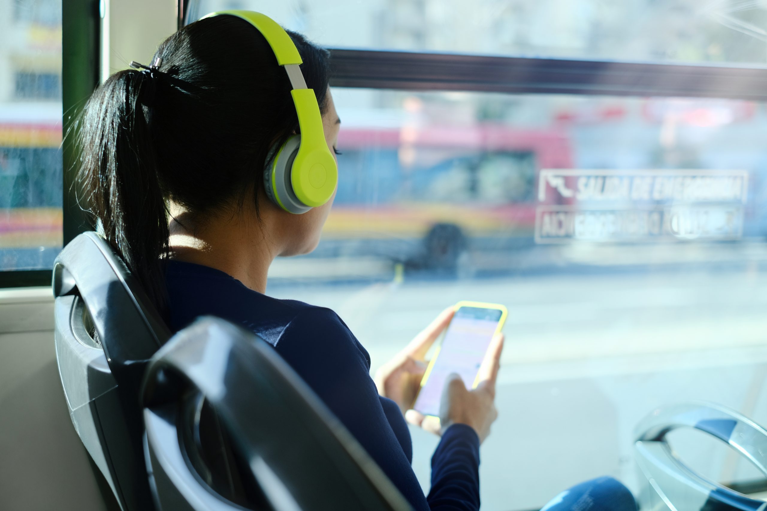 Woman With Headphones Listening To Music While Commuting To Work Using Public Transportation. Latin American Woman Sitting In A Bus And Using Mobile Phone.